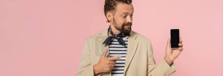 bearded french man in blazer and neck scarf pointing at smartphone with blank screen isolated on pink, banner