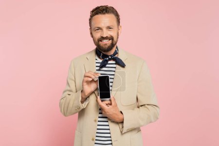heureux homme français en blazer et écharpe de cou tenant smartphone avec écran blanc isolé sur rose 