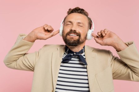 zufriedener französischer Mann in Blazer und Halstuch hört Musik in drahtlosen Kopfhörern isoliert auf rosa 