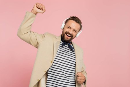 Photo for Excited french man in blazer and neck scarf listening music in wireless headphones isolated on pink - Royalty Free Image