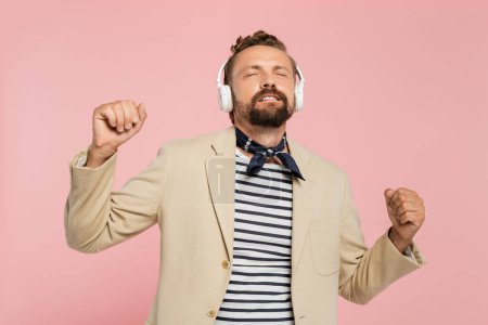 Foto de Hombre francés en chaqueta y cuello bufanda escuchar música en auriculares inalámbricos aislados en rosa - Imagen libre de derechos