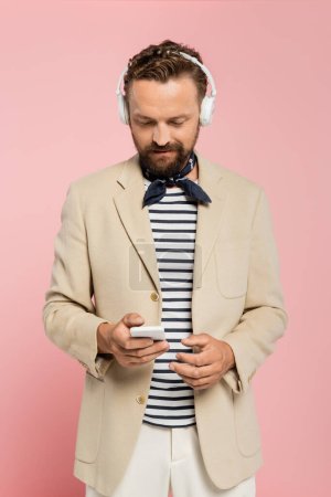 hombre francés barbudo en auriculares inalámbricos con teléfono inteligente aislado en rosa 
