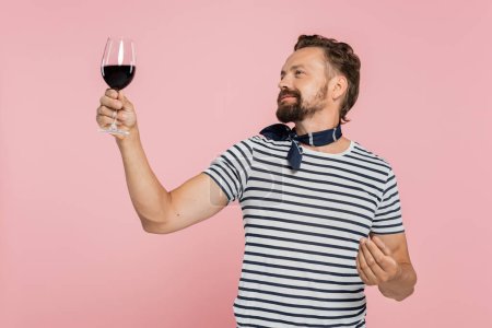 hombre alegre en camiseta rayada sosteniendo vaso de vino tinto francés aislado en rosa 