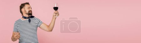 pleased man in striped t-shirt holding glass of red french wine isolated on pink, banner 