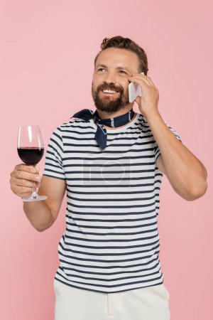 fröhlicher Mann im gestreiften T-Shirt, der ein Glas französischen Rotwein in der Hand hält und isoliert auf einem Smartphone in rosa spricht 