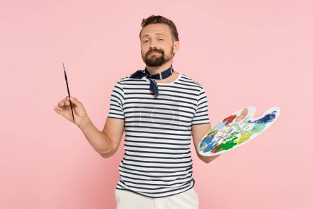 bearded french artist with neck scarf holding palette with colorful paints and paintbrush isolated on pink