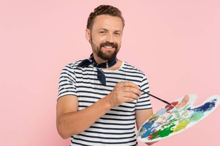 Téléchargez les photos : Artiste français positif avec écharpe cou tenant palette avec peintures colorées et pinceau isolé sur rose - en image libre de droit