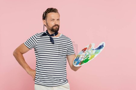 nachdenklicher französischer Künstler mit Halstuch, Palette mit bunten Farben isoliert auf rosa