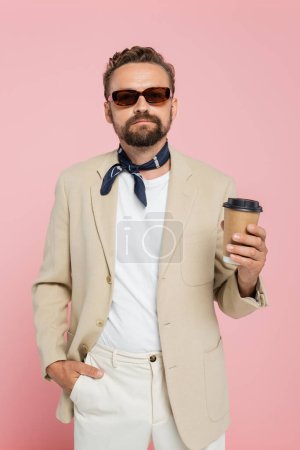 Foto de Elegante hombre francés en la bufanda del cuello y gafas de sol sosteniendo café para ir aislado en rosa - Imagen libre de derechos