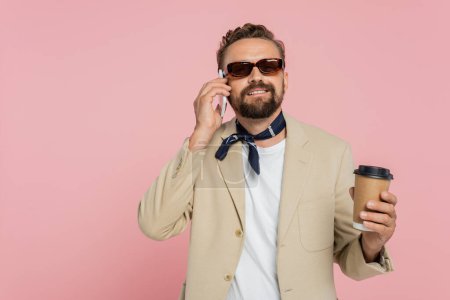 Téléchargez les photos : Heureux homme français dans le cou écharpe et lunettes de soleil tenant café pour aller et parler sur smartphone isolé sur rose - en image libre de droit