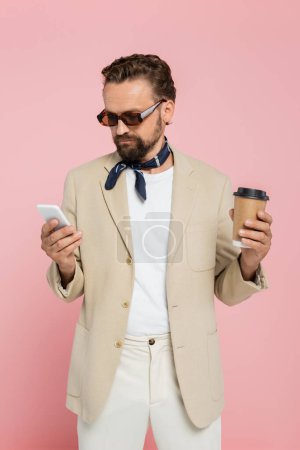 Foto de Elegante hombre francés en la bufanda del cuello y gafas de sol sosteniendo café para ir y el uso de teléfono inteligente aislado en rosa - Imagen libre de derechos