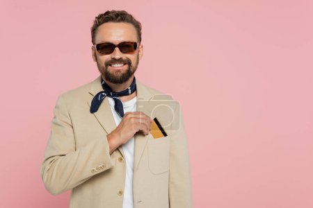 Téléchargez les photos : Homme heureux dans des lunettes de soleil élégantes et foulard de cou mettre carte de crédit dans la poche sur blazer isolé sur rose - en image libre de droit