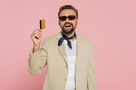 Téléchargez les photos : Homme excité dans des lunettes de soleil élégantes et foulard tenant la carte de crédit isolé sur rose - en image libre de droit