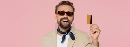 Téléchargez les photos : Homme excité dans des lunettes de soleil élégantes et foulard tenant la carte de crédit isolé sur rose, bannière - en image libre de droit