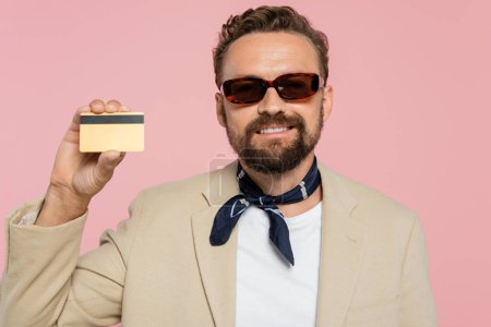 positive man in stylish sunglasses and neck scarf holding credit card isolated on pink 
