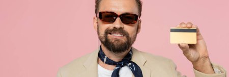 überglücklicher Mann mit stylischer Sonnenbrille und Halstuch, Kreditkarte isoliert auf rosa, Banner