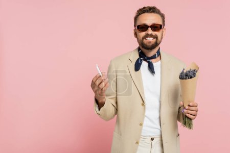 überglücklicher Mann in stylischer Sonnenbrille und Halstuch mit Blumen in Papierfolie und Smartphone isoliert auf rosa  