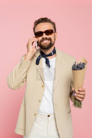 hombre feliz en gafas de sol con estilo y bufanda del cuello sosteniendo flores y hablando en el teléfono inteligente aislado en rosa  