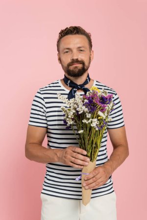 happy man in striped t-shirt and neck scarf holding flowers in paper wrap isolated on pink  