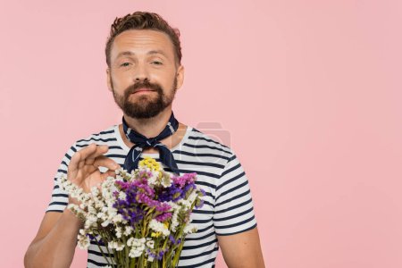 cheerful man in striped t-shirt and neck scarf holding wildflowers in paper wrap isolated on pink  