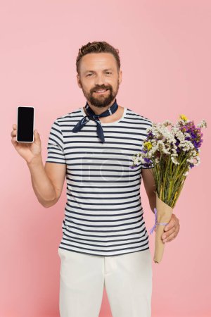 hombre alegre en camiseta a rayas y cuello bufanda celebración de flores silvestres y teléfono inteligente con pantalla en blanco aislado en rosa  