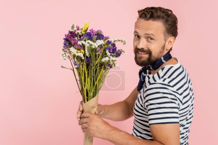 cheerful french man in striped t-shirt and neck scarf holding wildflowers isolated on pink  
