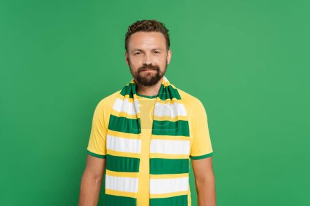 bearded sports fan in striped scarf and yellow t-shirt looking at camera isolated on green 