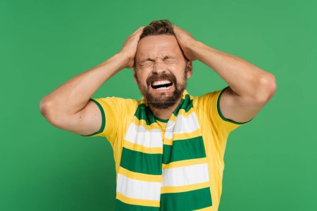 Photo for Emotional sports fan in striped scarf and yellow t-shirt pulling his hair isolated on green - Royalty Free Image