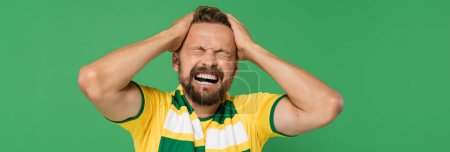Photo for Emotional sports fan in striped scarf and yellow t-shirt pulling his hair isolated on green, banner - Royalty Free Image