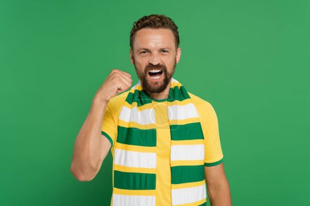 Emotionaler Sportfan in gestreiftem Schal und gelbem T-Shirt blickt in die Kamera und zeigt Machtgeste isoliert auf Grün 