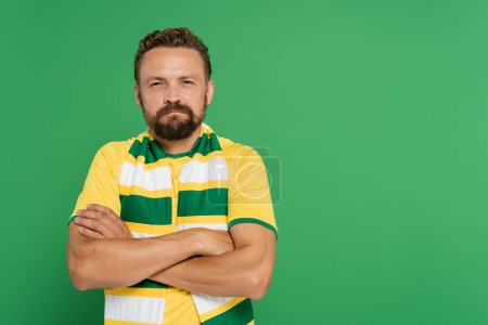 Foto de Abanico de fútbol ofendido en bufanda a rayas y camiseta amarilla de pie con los brazos cruzados aislados en verde - Imagen libre de derechos