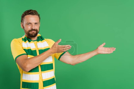 Photo for Bearded soccer fan in striped scarf and yellow t-shirt looking at camera while pointing with hands isolated on green - Royalty Free Image