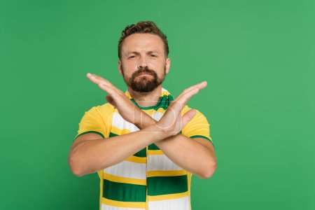 bearded soccer fan in striped scarf and yellow t-shirt showing stop gesture with hands isolated on green 