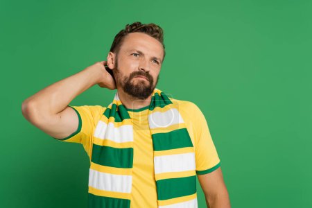 Nachdenklicher Fußballfan in gestreiftem Schal und gelbem T-Shirt, der isoliert auf Grün blickt 