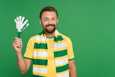 alegre ventilador de fútbol en bufanda a rayas sosteniendo aplausos de mano de plástico aislado en verde 