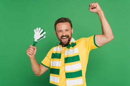 Photo for Excited football fan in striped scarf holding plastic hand clapper isolated on green - Royalty Free Image