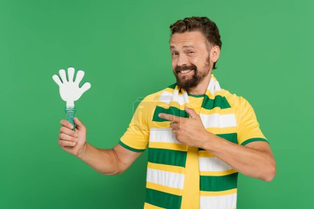 cheerful football fan in striped scarf pointing with finger at plastic hand clapper isolated on green 