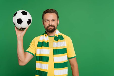 bärtiger Fußballfan in gestreiftem Schal und gelbem T-Shirt, der Fußball isoliert auf Grün hält 