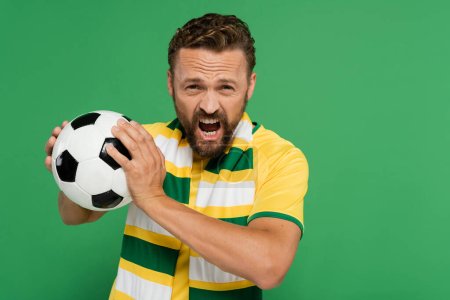 emotional soccer fan in striped scarf and yellow t-shirt holding football isolated on green 