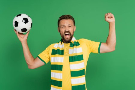 Emotionaler Fußballfan mit gestreiftem Schal hält Fußball in der Hand und jubelt isoliert auf Grün 
