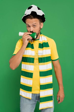 bearded man in sports hat blowing horn while cheering isolated on green 