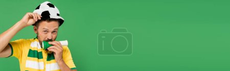 Téléchargez les photos : Homme barbu dans le chapeau de football souffle corne tout en acclamant isolé sur vert, bannière - en image libre de droit