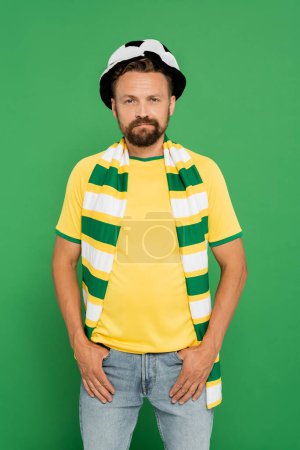 bearded man in sports fan hat and striped scarf standing with hands in pockets isolated on green 