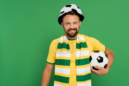 glücklicher bärtiger Mann mit Fußballfan-Mütze und gestreiftem Schal, der mit Ball isoliert auf Grün steht 