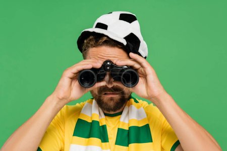 bearded man in football fan hat and striped scarf looking through binoculars during match isolated on green 