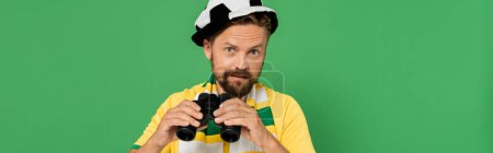 bearded man in fan hat and striped scarf holding binoculars during football match isolated on green, banner 