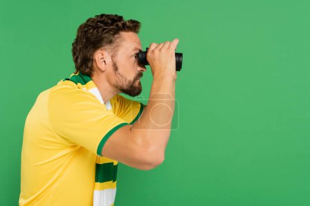 Seitenansicht eines bärtigen Mannes in gestreiftem Schal mit Fernglas während eines Fußballspiels isoliert auf grün