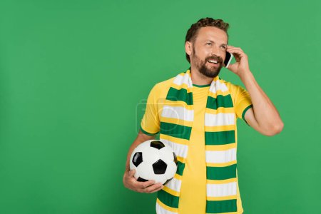 fröhlicher Mann mit gestreiftem Schal hält Fußball in der Hand und telefoniert isoliert auf Grün