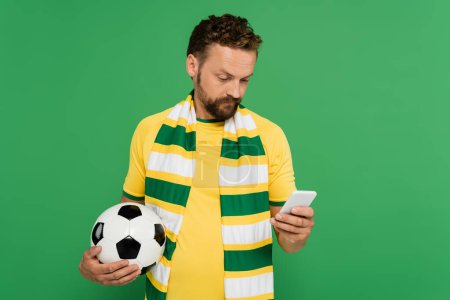 hombre barbudo en bufanda a rayas que sostiene el fútbol y el uso de teléfono móvil aislado en verde