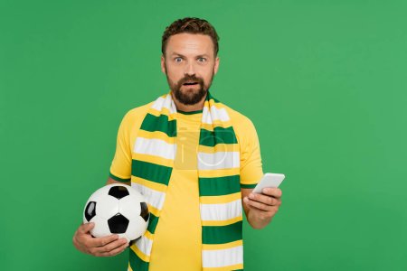 Betäubter Mann mit gestreiftem Schal hält Fußball und Handy isoliert auf Grün